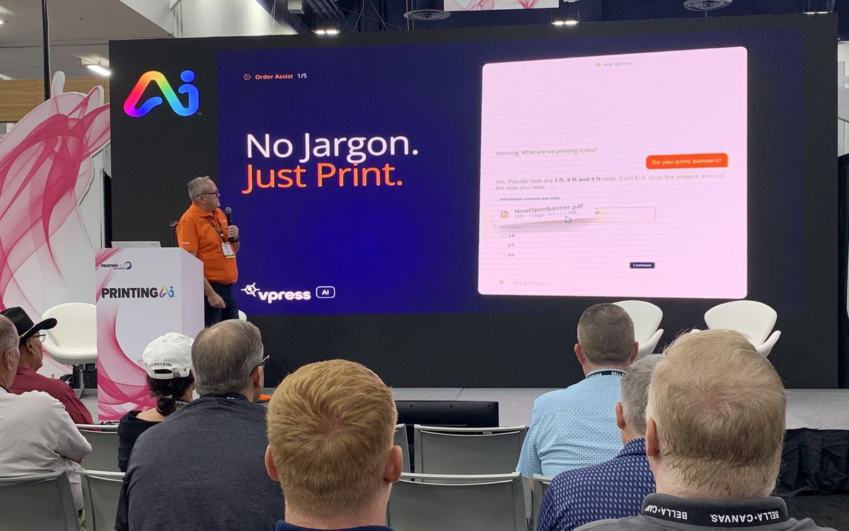 Kelvin Bell presenting Order Assist on stage, with a slide reading No Jargon. Just Print.